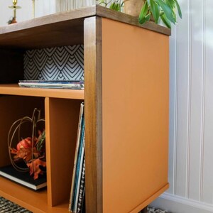 May include: A mid-century modern style record player stand with a burnt orange finish and wood trim. The stand has three shelves and is decorated with a small pumpkin and fall leaves.