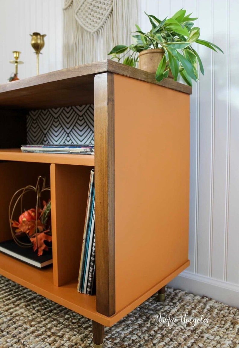 May include: A mid-century modern style record player stand with a burnt orange finish and wood trim. The stand has three shelves and is decorated with a small pumpkin and fall leaves.
