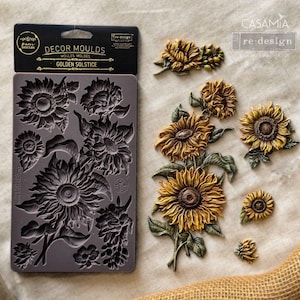 May include: A grey silicone decor mould labeled Golden Solstice with sunflower designs, accompanied by several finished sunflower castings in yellow, brown, and green. The packaging features the text Decor Moulds and re-design with prima.