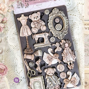 May include: A dark gray silicone mold featuring various embossed shapes including a dress form, a teddy bear, an ornate oval frame, a sewing machine, a swan, a doll, scissors, a spool of thread, a book, and several small circular buttons.