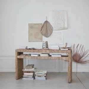 May include: A natural woven console table with two shelves, featuring a light brown finish. The table is decorated with stacks of books and a decorative palm leaf.