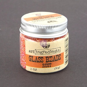 May include: A clear plastic jar filled with small, iridescent rust-colored glass beads. The jar features a silver screw-top lid and a tan label with the text art ingredients GLASS BEADS RUST 2 OZ (56 g) and a butterfly logo.