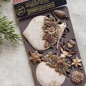 May include: A brown silicone mold for crafting, labeled DECOR MOULDS EVERGREEN ANTLER EMBLEM. The mold features cavities for casting two large circular ornaments with pinecone and floral details, a bird, a deer head, an acorn, a star, and a pinecone.