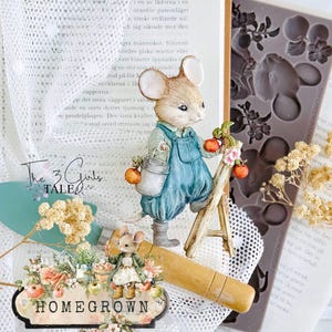 May include: A whimsical figurine of a mouse in blue overalls, holding a bucket and climbing a small ladder to pick apples. The scene includes a "HOMEGROWN" sign, dried flowers, and a book. A gardening theme.