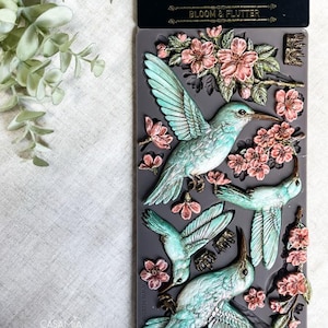May include: A gray silicone mold featuring hummingbird and floral designs. The hummingbirds are teal and white with detailed wings, and the flowers are pink. The mold is labeled "DECOR MOULDS BLOOM & FLUTTER".