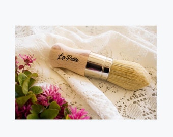 May include: A wooden-handled paint brush with a natural bristle head. The handle is labeled "La Petite" in black lettering. The brush is lying on a white lace fabric.