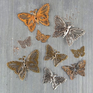 May include: Assortment of metal butterfly charms in various sizes and finishes, including silver, gold, and copper. Some butterflies have a distressed look, while others are shiny. The butterflies are arranged on a gray background.