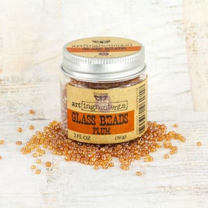 May include: A clear plastic jar with a silver metal lid containing small, translucent amber-colored glass beads. The label reads Art Ingredients Glass Beads Plum, 2 fl oz. A pile of the loose beads is scattered around the base of the jar.