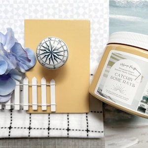 May include: A decorative compass rose knob with a white and navy blue design is mounted on a yellow background. A jar of paint with the text "CATCHIN' SOME RAYS" is next to the knob. A small white picket fence and blue flowers are also in the image.