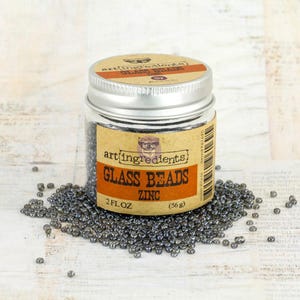 May include: A clear plastic jar with a silver metal lid containing small, dark metallic gray glass beads. The label reads Art Ingredients Glass Beads Zinc 2 FL OZ (56 g). A pile of the same dark gray beads is scattered around the base of the jar.
