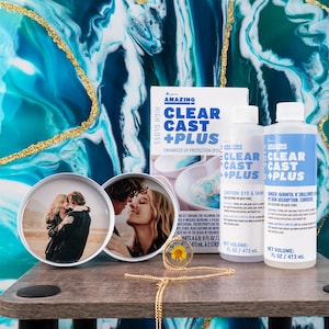 May include: A display of epoxy resin supplies and finished resin art. The kit includes bottles of "CLEAR CAST + PLUS" epoxy, two resin-covered photo coasters, and a sunflower pendant on a gold chain. The background features a blue and gold abstract design.