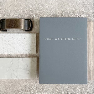 May include: A gray sample board with the text "GONE WITH THE GRAY" on it. The board is next to two white countertop samples.
