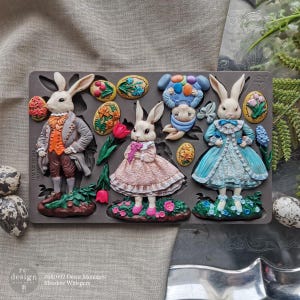 May include: A gray silicone mold featuring Easter-themed designs. The mold includes detailed bunny figures in Victorian-style clothing, Easter eggs, and floral accents. The text "#681692 Decor Moulds Meadow Whispers" is visible.