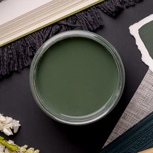 May include: A small glass jar filled with a dark green paint. The jar is sitting on a black surface with a book and a piece of paper in the background.