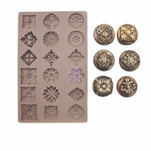 May include: A silicone mold with various floral and geometric designs, alongside several finished, decorative buttons. The mold features detailed patterns, including flowers, rosettes, and symmetrical shapes. The buttons have a metallic finish.