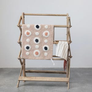 May include: A wooden drying rack with three tiers, featuring a pink and white rug with black and orange circles hanging on the top tier. A white and black striped towel hangs on the middle tier.