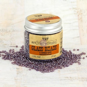 May include: A clear glass jar with a silver metal lid containing small, dark purple glass beads. The label reads Art Ingredients Glass Beads Licorice 2 fl oz (56 g). A pile of the same dark purple beads is scattered around the base of the jar.