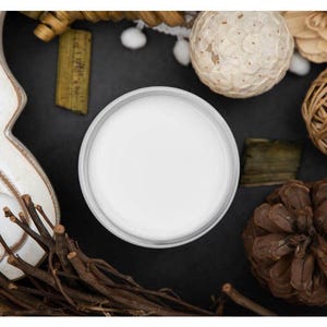 May include: A white candle in a glass jar on a black surface surrounded by decorative items, including a pine cone, woven balls, and a piece of dried foliage.