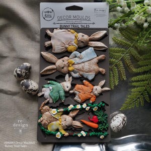 May include: A dark gray silicone mold featuring detailed bunny designs in various outfits, including jackets and dresses. The mold is labeled "DECOR MOULDS BUNNY TRAIL TALES" and includes two speckled eggs. The overall theme is Easter and spring.