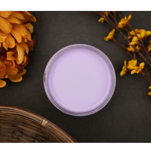 May include: A small glass jar filled with a light purple cream or lotion. The jar is sitting on a dark gray surface.