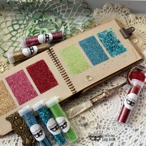 May include: A collection of small glass jars filled with different colors of glitter. The jars are arranged in a row on a brown notebook with pages of glitter swatches. The notebook is open and the pages are fanned out. The glitter swatches are in the colors gold, pink, red, green, blue, and turquoise. The jars are labeled with the words "Finnabair".