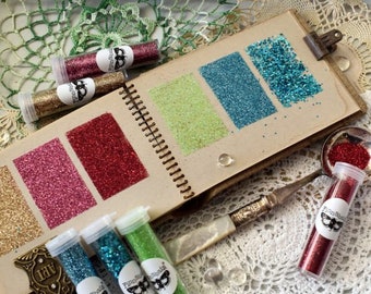 May include: A collection of small glass jars filled with different colors of glitter. The jars are arranged in a row on a brown notebook with pages of glitter swatches. The notebook is open and the pages are fanned out. The glitter swatches are in the colors gold, pink, red, green, blue, and turquoise. The jars are labeled with the words "Finnabair".
