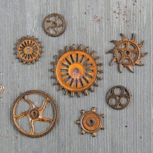 May include: A collection of eight rusty metal gears in various sizes and shapes, with a gray wooden background. The gears are all in shades of brown and orange, with some showing signs of wear and tear.
