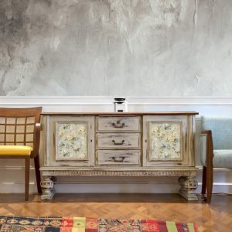 Puede incluir: Un aparador desgastado de color blanquecino con tres cajones y paneles florales. El aparador tiene una tapa de madera y se asienta sobre patas ornamentadas. A la izquierda hay una silla de madera con asiento amarillo y a la derecha un sof&aacute; azul.