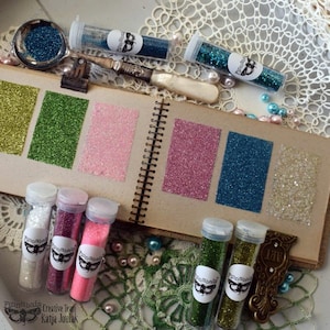 May include: A collection of small glass bottles filled with various colors of glitter. The bottles are labeled with the text "Finnabair Creative Team Katja Joulak". The glitter is displayed on a page in a scrapbook with a variety of colors, including green, pink, blue, and white.