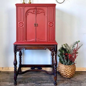 Pu&ograve; includere: Un mobile vintage rosso con una base in legno e dettagli decorativi. Il mobile ha due ante e uno scaffale all'interno. Il mobile &egrave; posizionato su un pavimento in legno.