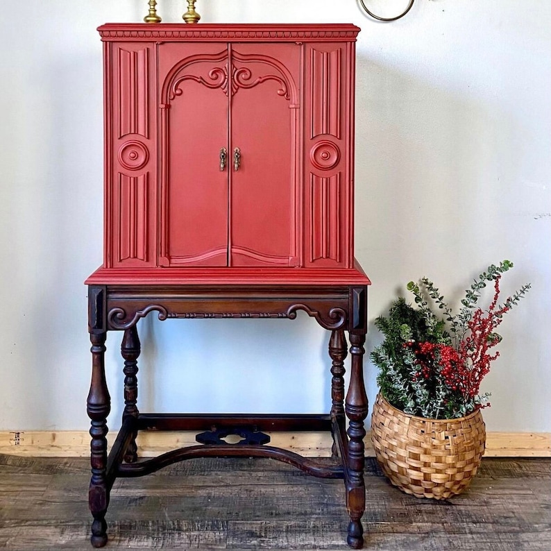 Pu&ograve; includere: Un mobile vintage rosso con una base in legno e dettagli decorativi. Il mobile ha due ante e uno scaffale all'interno. Il mobile &egrave; posizionato su un pavimento in legno.