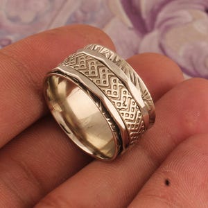 May include: A silver-colored ring with intricate geometric patterns. The ring features a textured design around the band, with a central rotating element. The ring is shown in a close-up shot, highlighting the detailed craftsmanship.