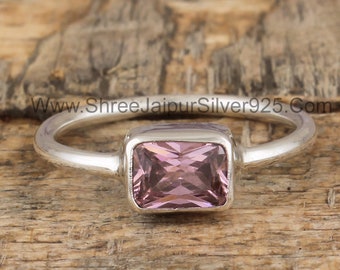 May include: A silver ring with a light pink square-shaped gemstone set in a silver bezel. The ring is on a wooden surface.