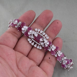 Pode incluir: Uma pulseira prateada com uma pedra central retangular de cor rubi, rodeada por pedras brancas e de cor rubi menores num padrão floral. A pulseira é segurada em uma mão, mostrando seu design intrincado e detalhes brilhantes.