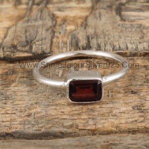 May include: A silver ring with a rectangular, dark red gemstone. The ring is set on a wooden surface. www.shreejaipursilver925.com