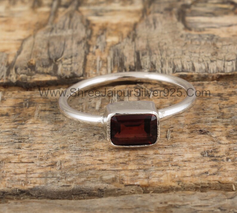 May include: A silver ring with a rectangular, dark red gemstone. The ring is set on a wooden surface. www.shreejaipursilver925.com
