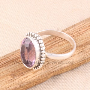 Pu&ograve; includere: Un anello d'argento con una grande pietra di ametista ovale, viola chiaro. La gemma &egrave; circondata da un bordo perlato. L'anello &egrave; appoggiato su una superficie di legno. Il sito web "www.ShreeJaipurSilver925.com" &egrave; visibile.