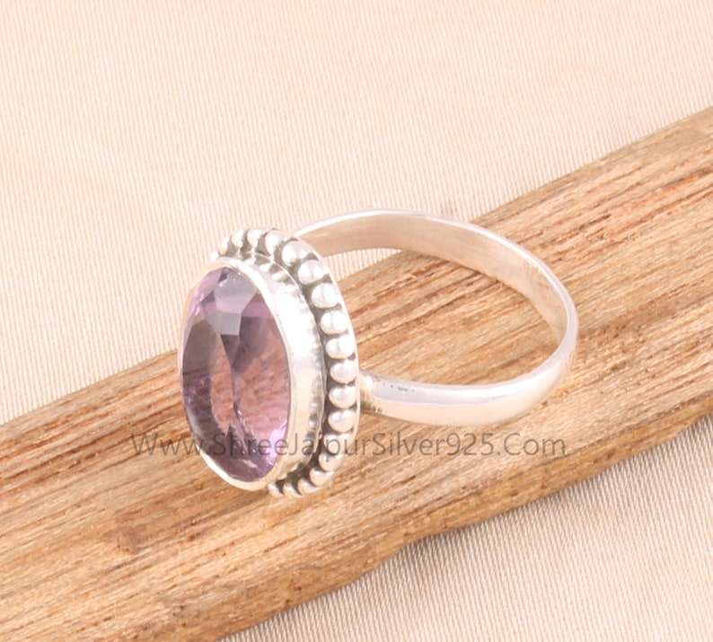 Pu&ograve; includere: Un anello d'argento con una grande pietra di ametista ovale, viola chiaro. La gemma &egrave; circondata da un bordo perlato. L'anello &egrave; appoggiato su una superficie di legno. Il sito web "www.ShreeJaipurSilver925.com" &egrave; visibile.