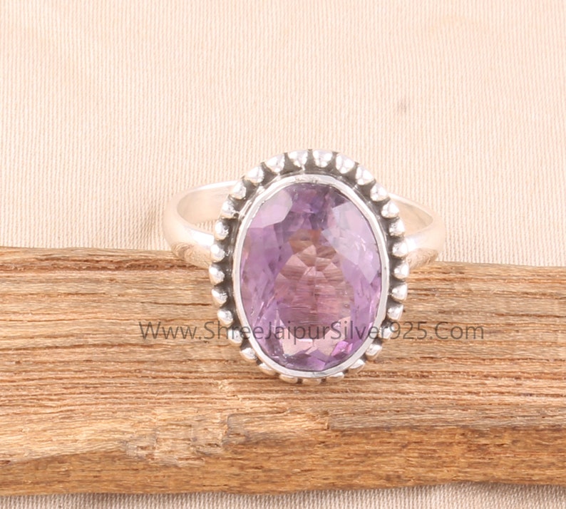 Pu&ograve; includere: Un anello d'argento con una grande ametista ovale. La gemma, di colore viola chiaro, &egrave; incastonata in una fascia d'argento con un bordo di perline. L'anello &egrave; esposto su una superficie di legno.