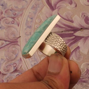 Op de afbeelding: Een zilveren ring met een grote, rechthoekige, lichtblauwe edelsteen. De band heeft een textuur met stippen. De ring wordt omhoog gehouden, waarbij de kleur van de edelsteen en de ingewikkelde details van de band worden benadrukt.