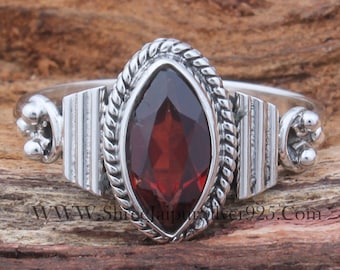 Puede incluir: Un anillo de plata con una piedra preciosa roja engastada en un diseño de cuerda trenzada. El anillo tiene una banda texturizada con un detalle decorativo a cada lado.