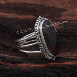 May include: A silver ring featuring a dark red oval gemstone set in a silver bezel. The ring has a triple band design and a beaded edge around the bezel. The website address "WWW.ShreeGaurSilver925.Com" is visible in the background.