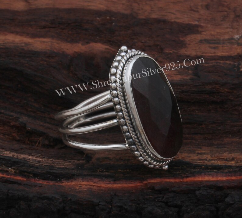 May include: A silver ring featuring a dark red oval gemstone set in a silver bezel. The ring has a triple band design and a beaded edge around the bezel. The website address "WWW.ShreeGaurSilver925.Com" is visible in the background.