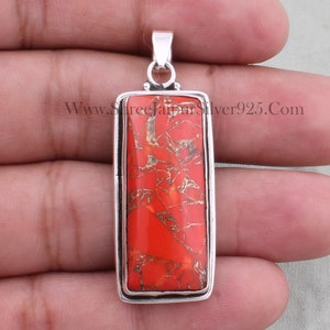 May include: A silver pendant featuring a rectangular red coral stone. The stone has a marbled pattern with white and gold veins. The pendant is displayed on a hand.