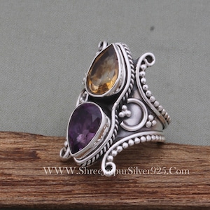 May include: A silver ring with a purple and yellow gemstone setting. The ring has a detailed design with intricate swirls and beading.