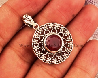 May include: A silver pendant featuring a red gemstone set within a filigree design. The pendant is round and has a delicate floral pattern.