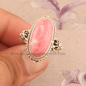 May include: A silver ring featuring a pink oval gemstone set within a floral design. The ring is worn on a finger.
