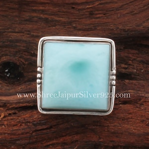 May include: A square-shaped ring with a light blue gemstone set in a silver frame. The ring is on a wooden surface.
