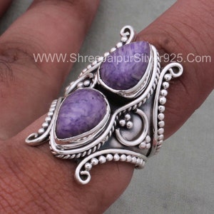 May include: A silver ring with two purple stones set in a decorative design. The ring is wide and has a textured band. The stones are a light purple color and have a swirling pattern. The ring is on a finger.