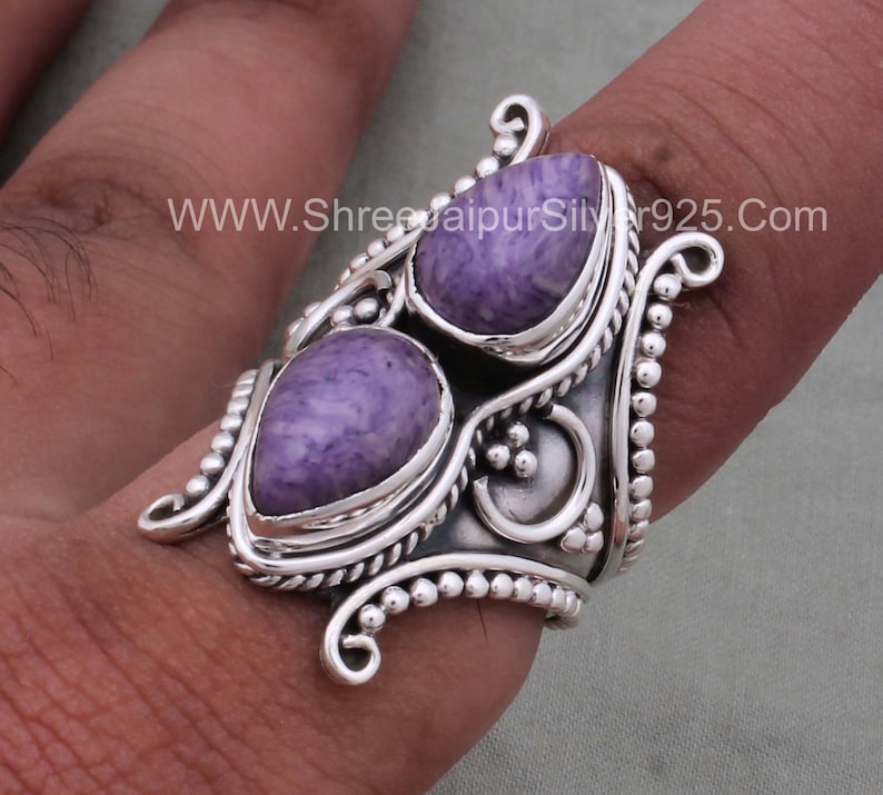 May include: A silver ring with two purple stones set in a decorative design. The ring is wide and has a textured band. The stones are a light purple color and have a swirling pattern. The ring is on a finger.
